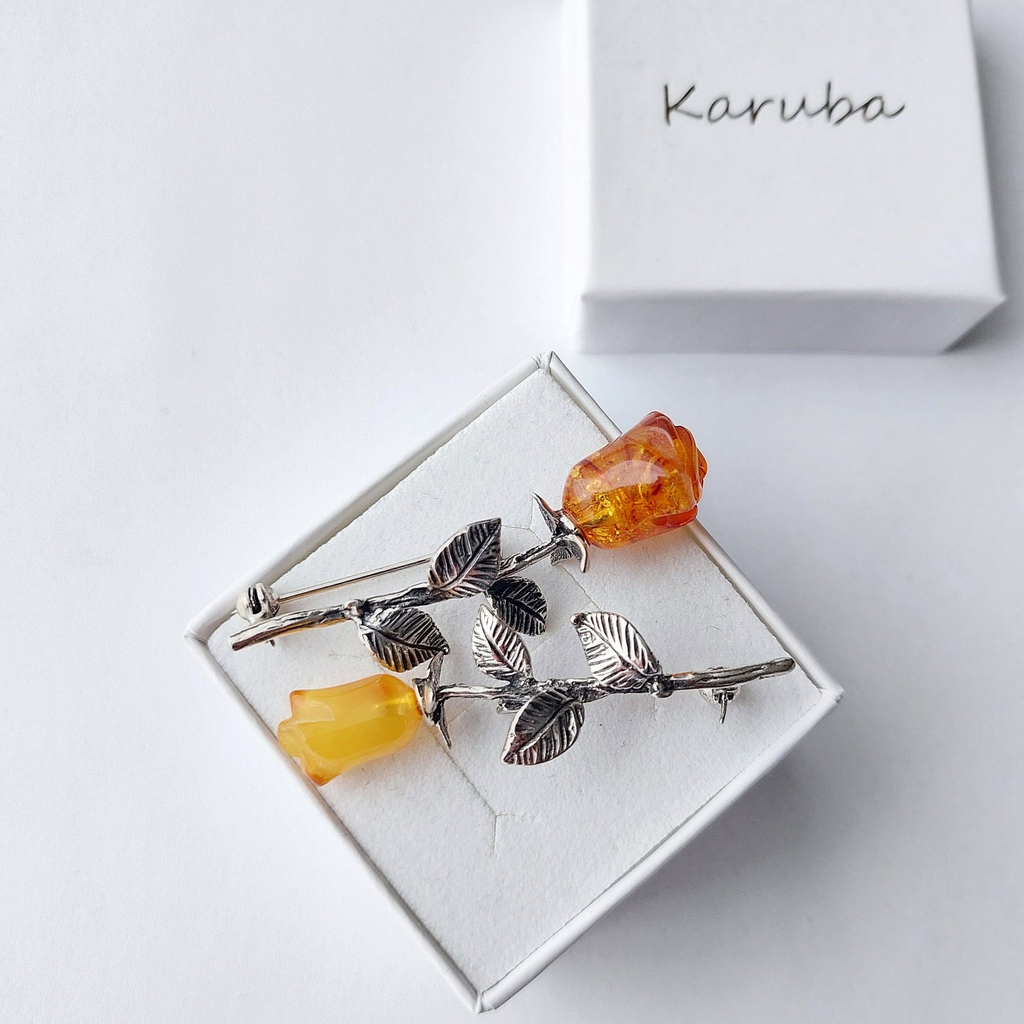 Silver brooch with amber stones on a white box, branded 'Karuba'.