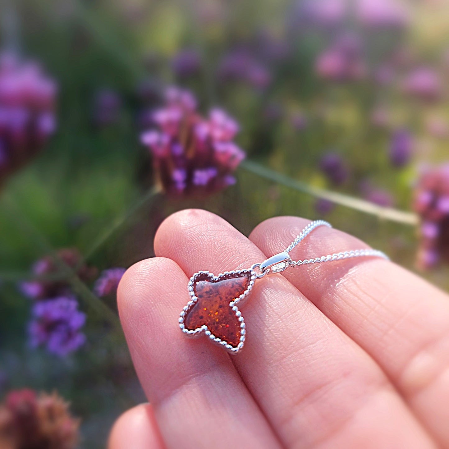 Butterfly-shaped pendant on a chain held against a blurred natural background with purple flowers.