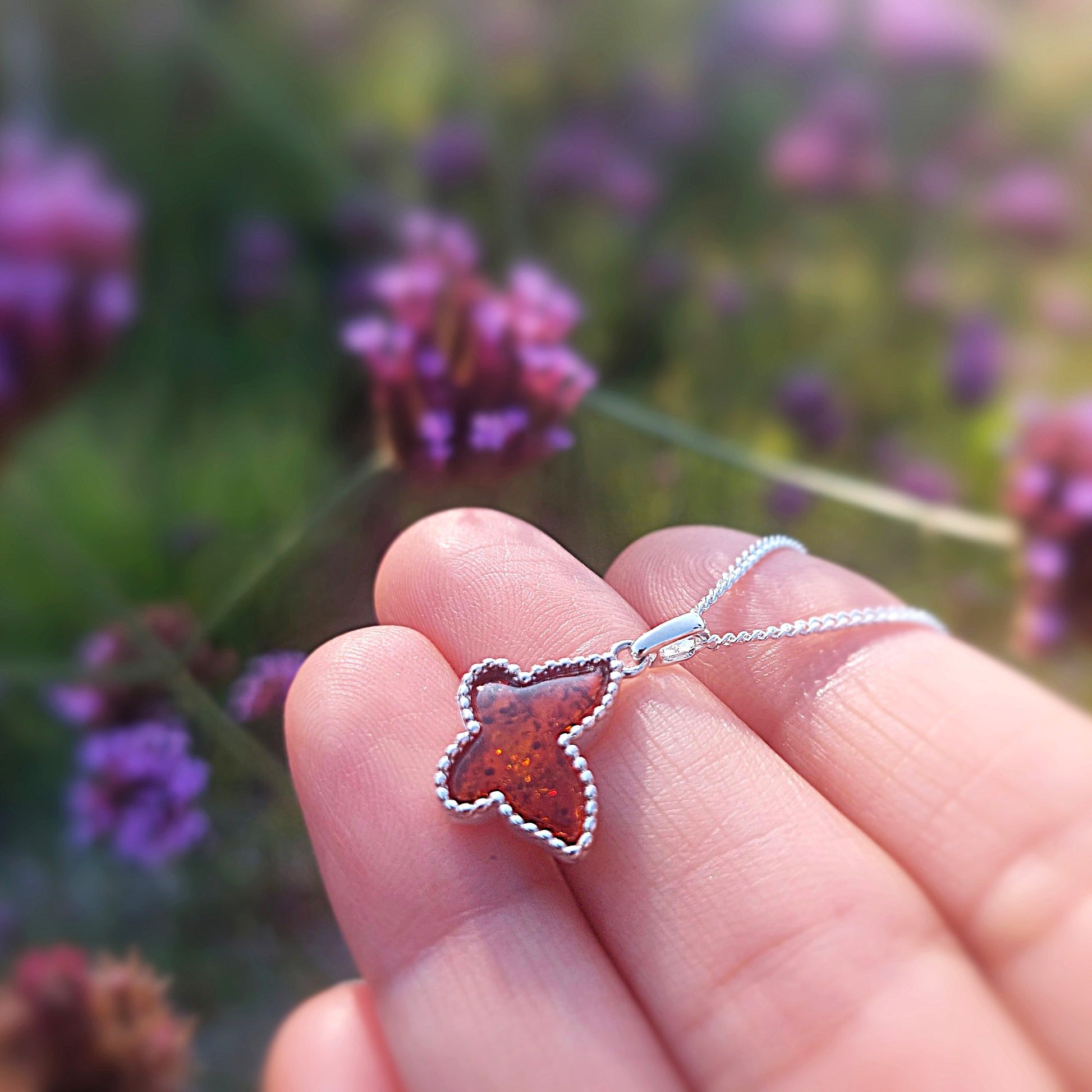 Butterfly-shaped pendant on a chain held against a blurred natural background with purple flowers.
