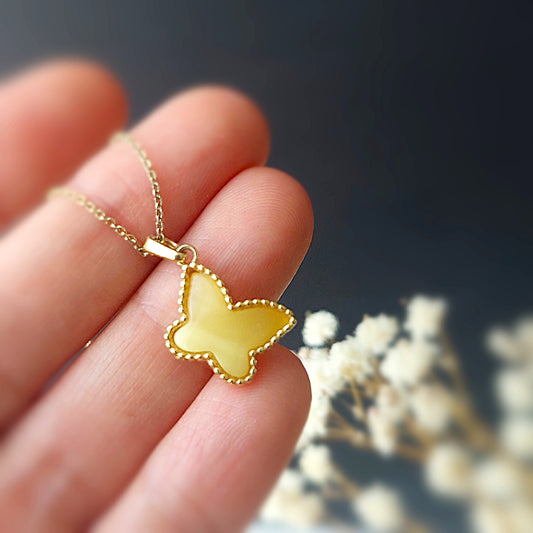 Yellow butterfly pendant on a gold chain held between fingers with a blurred floral background