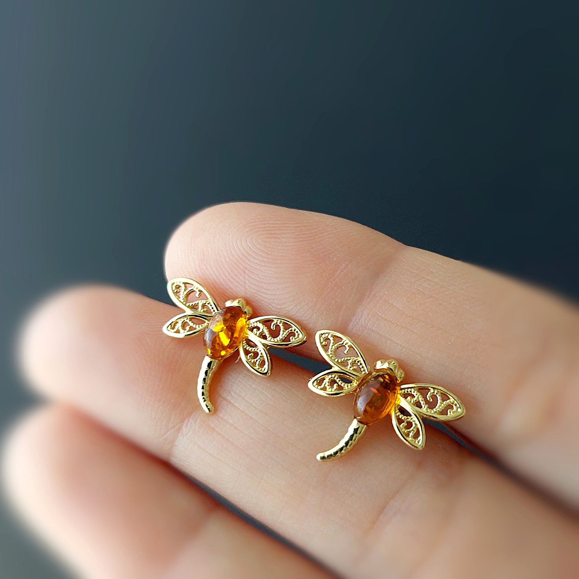 Gold dragonfly earrings with amber stones held between fingers against a dark background