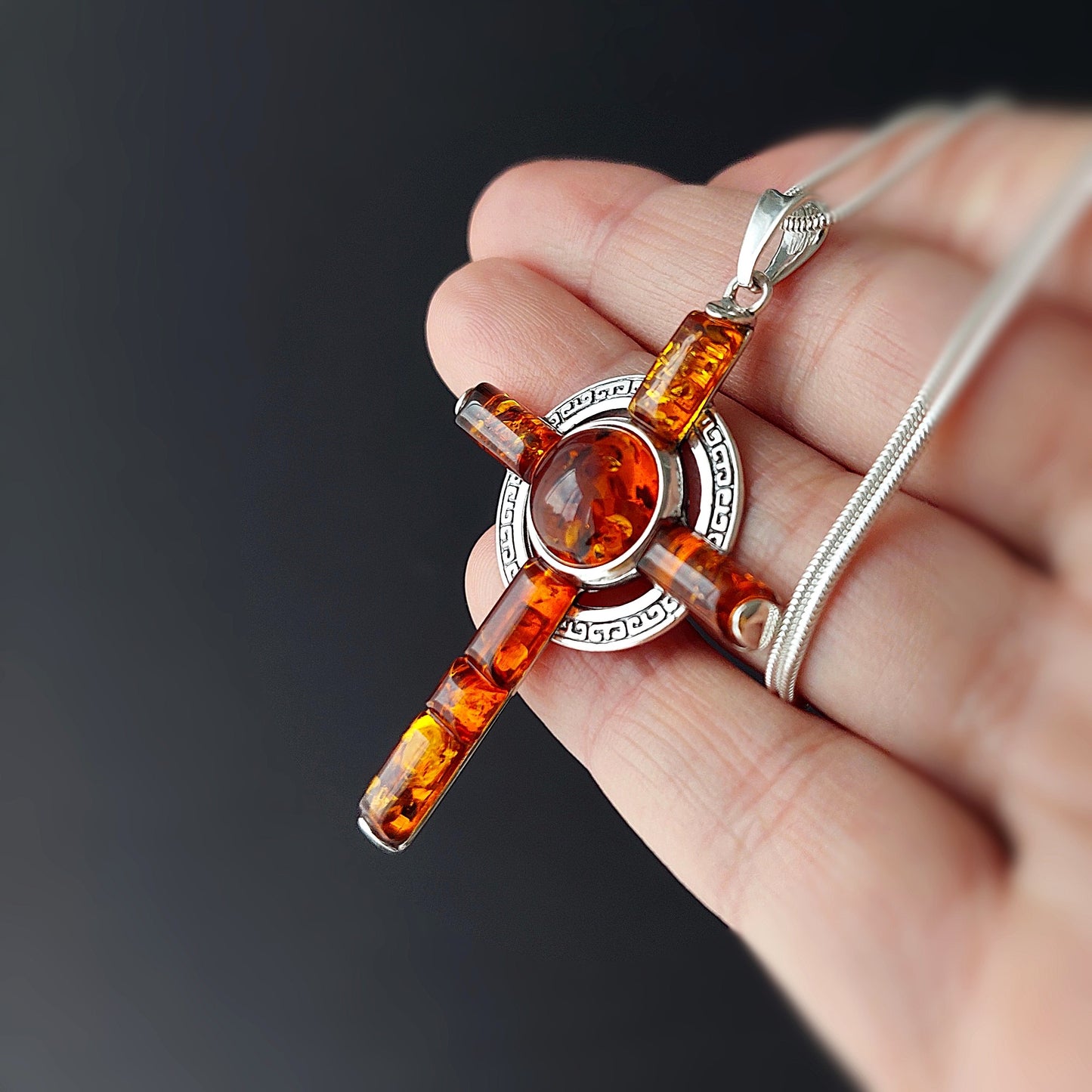 Amber cross pendant on a chain held in a hand against a dark background